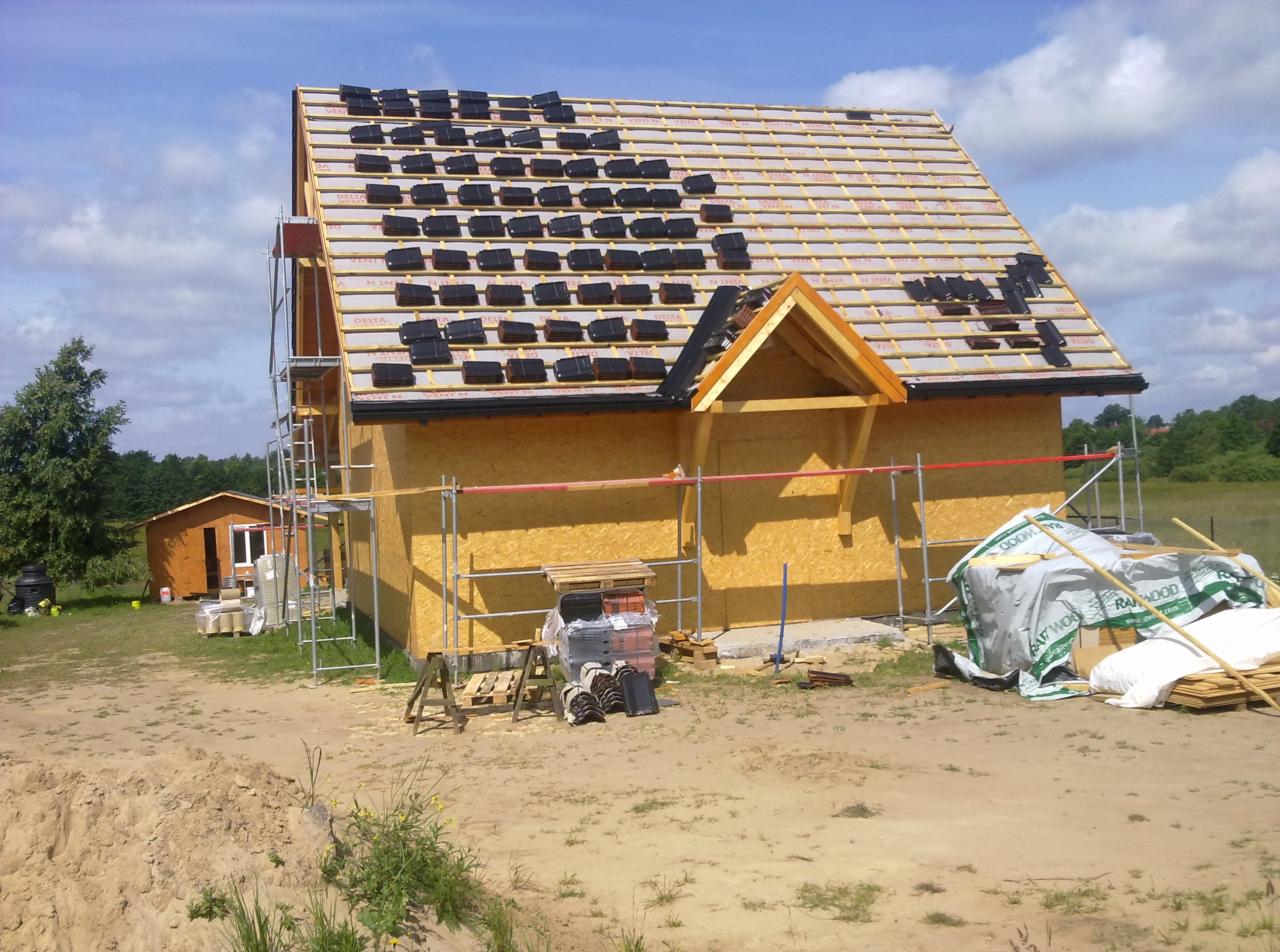 Wznoszenie domu w technologii szkieletowej: montaż dachu z czarnej dachówki ceramicznej, widoczne rusztowania i materiały budowlane na placu budowy.