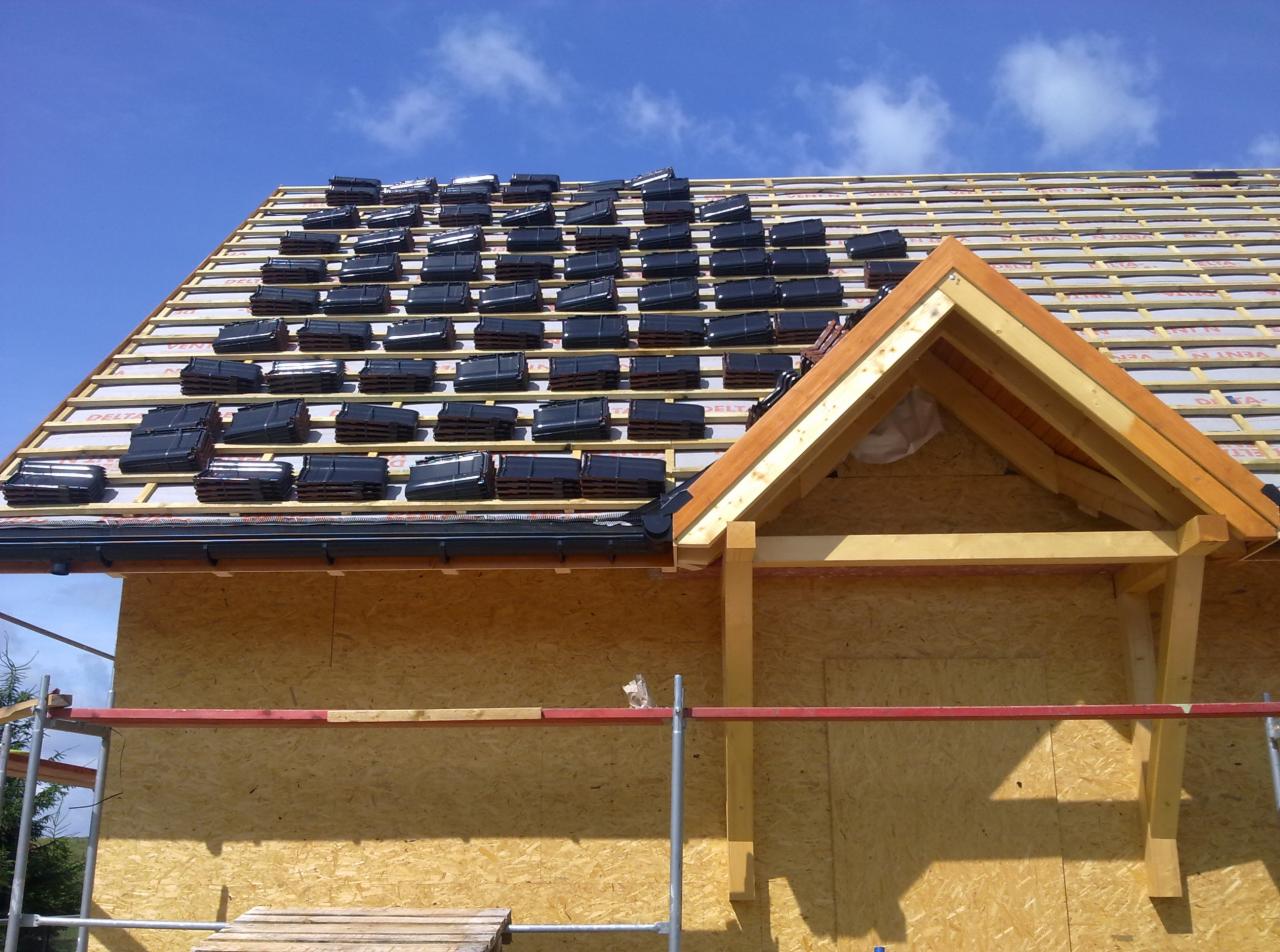 Fragment dachu w trakcie krycia dachówką ceramiczną, widoczny drewniany szkielet konstrukcji, membrany i łaty, z bliska ułożone rzędy dachówek na połaci dachu pod błękitnym niebem.