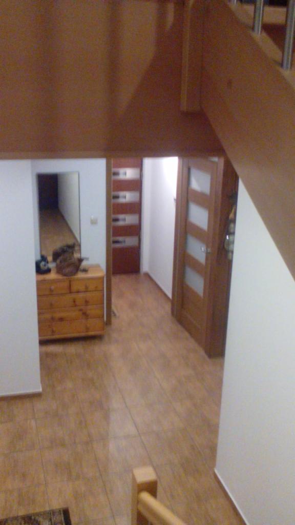 Wnętrze domu z widokiem na korytarz z drewnianą podłogą i komodą z lustrem, widoczne drewniane schody z balustradą.