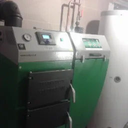 Zielono-szary piec na paliwo stałe marki Termo-Tech Stąporków o mocy 15 kW z podłączonym kominem i zasobnikiem ciepłej wody w kotłowni wyłożonej płytkami.