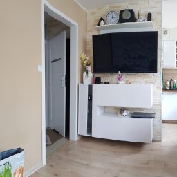 Remonty A.R.Z - Jasne wnętrze po remoncie: ściana z imitacji kamienia, biała szafka RTV z dekoracjami, telewizor, drewniana podłoga, widok na kuchnię i otwarte drzwi.
