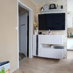 Jasne wnętrze po remoncie: ściana z imitacji kamienia, biała szafka RTV z dekoracjami, telewizor, drewniana podłoga, widok na kuchnię i otwarte drzwi.