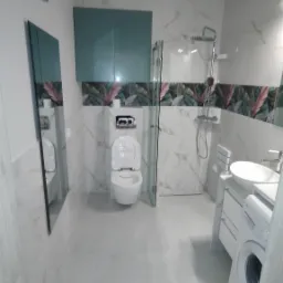 Nowoczesna łazienka z kabiną prysznicową, toaletą i pralką. Białe płytki imitujące marmur, szafki w odcieniach zieleni i motyw roślinny na dekorze ściennym.