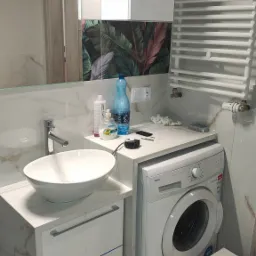 Nowoczesna łazienka z umywalką nablatową, marmurowymi płytkami i motywem tropikalnych liści na ścianie. Biała pralka i grzejnik drabinkowy.