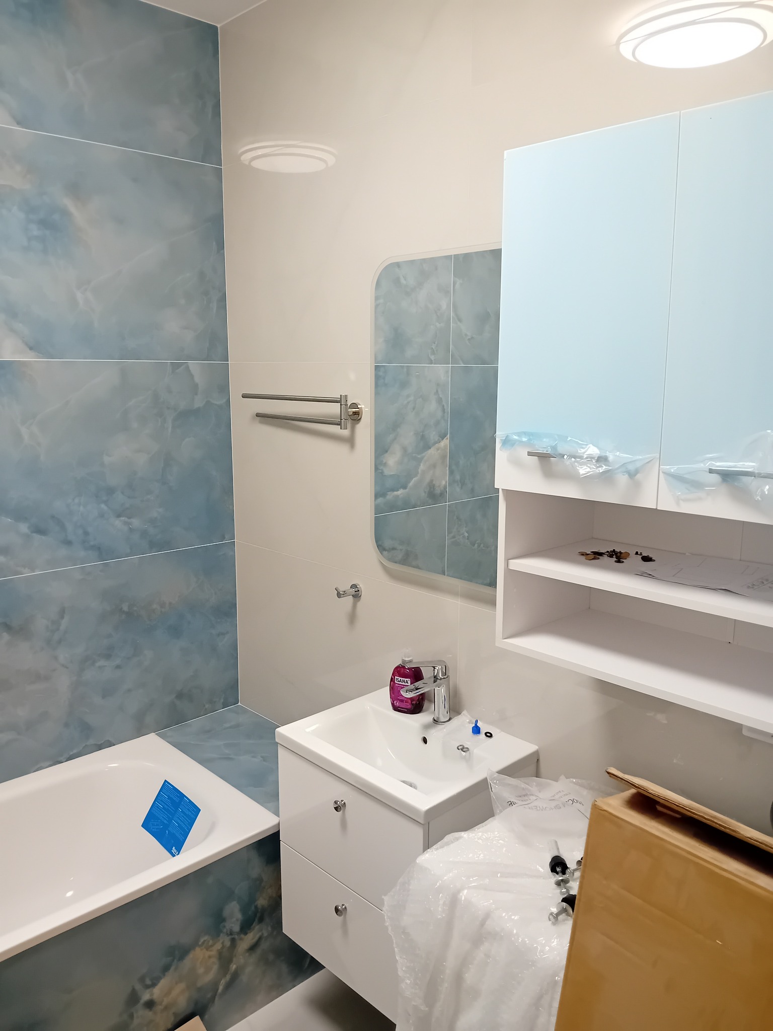 Niebiesko-biała łazienka z marmurkowymi płytkami, białą armaturą i szafką z błękitnymi frontami, częściowo jeszcze zabezpieczonymi folią, w trakcie montażu, widoczne elementy montażowe i kartonowe...