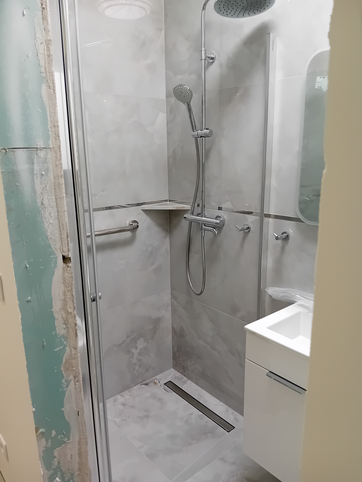 Wnętrze łazienki w trakcie prac: kabina prysznicowa z chromowaną armaturą, płytki imitujące marmur, umywalka z szafką, widoczne niedokończone ściany przed położeniem płytek.