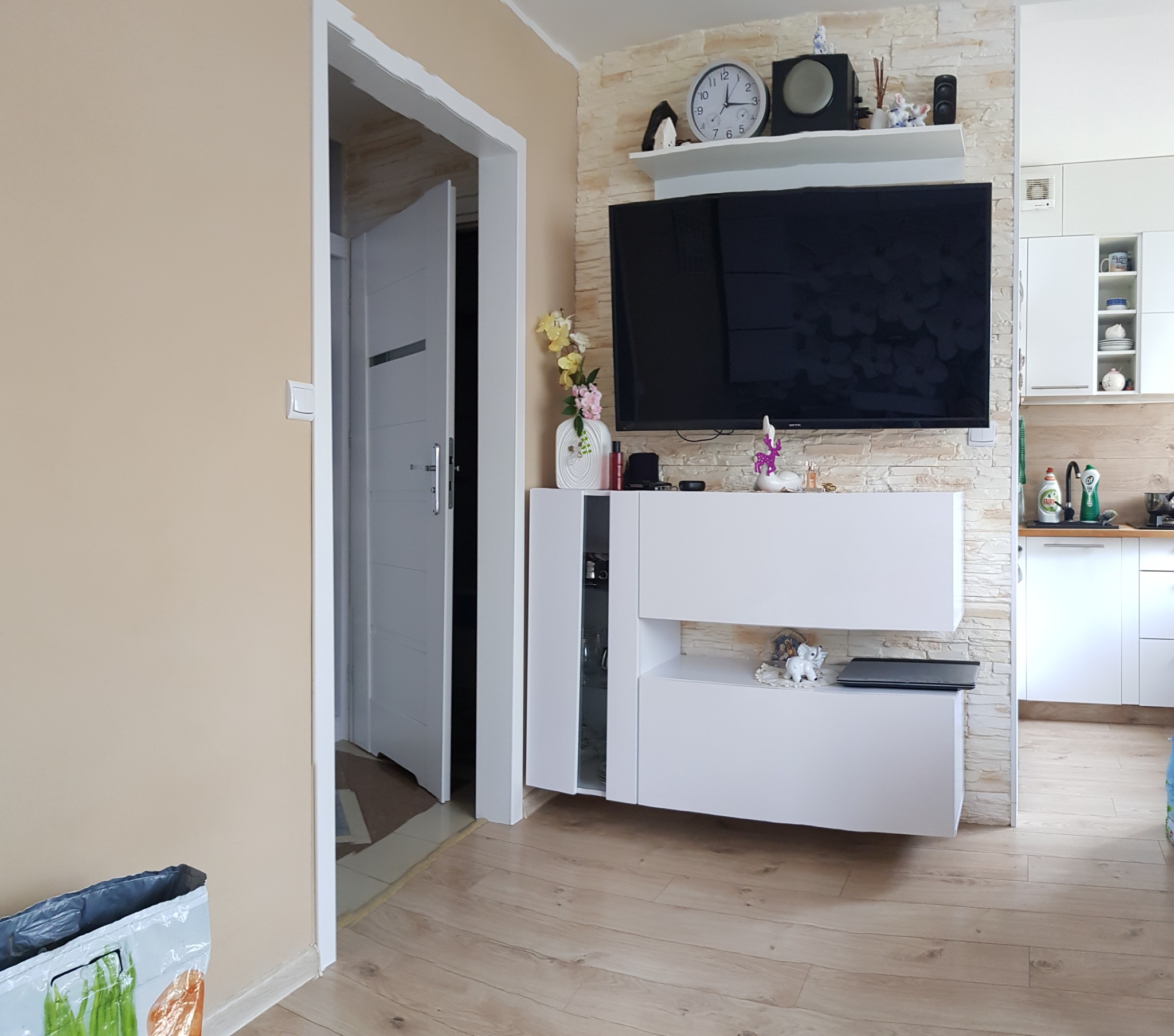 Jasne wnętrze po remoncie: ściana z imitacji kamienia, biała szafka RTV z dekoracjami, telewizor, drewniana podłoga, widok na kuchnię i otwarte drzwi.