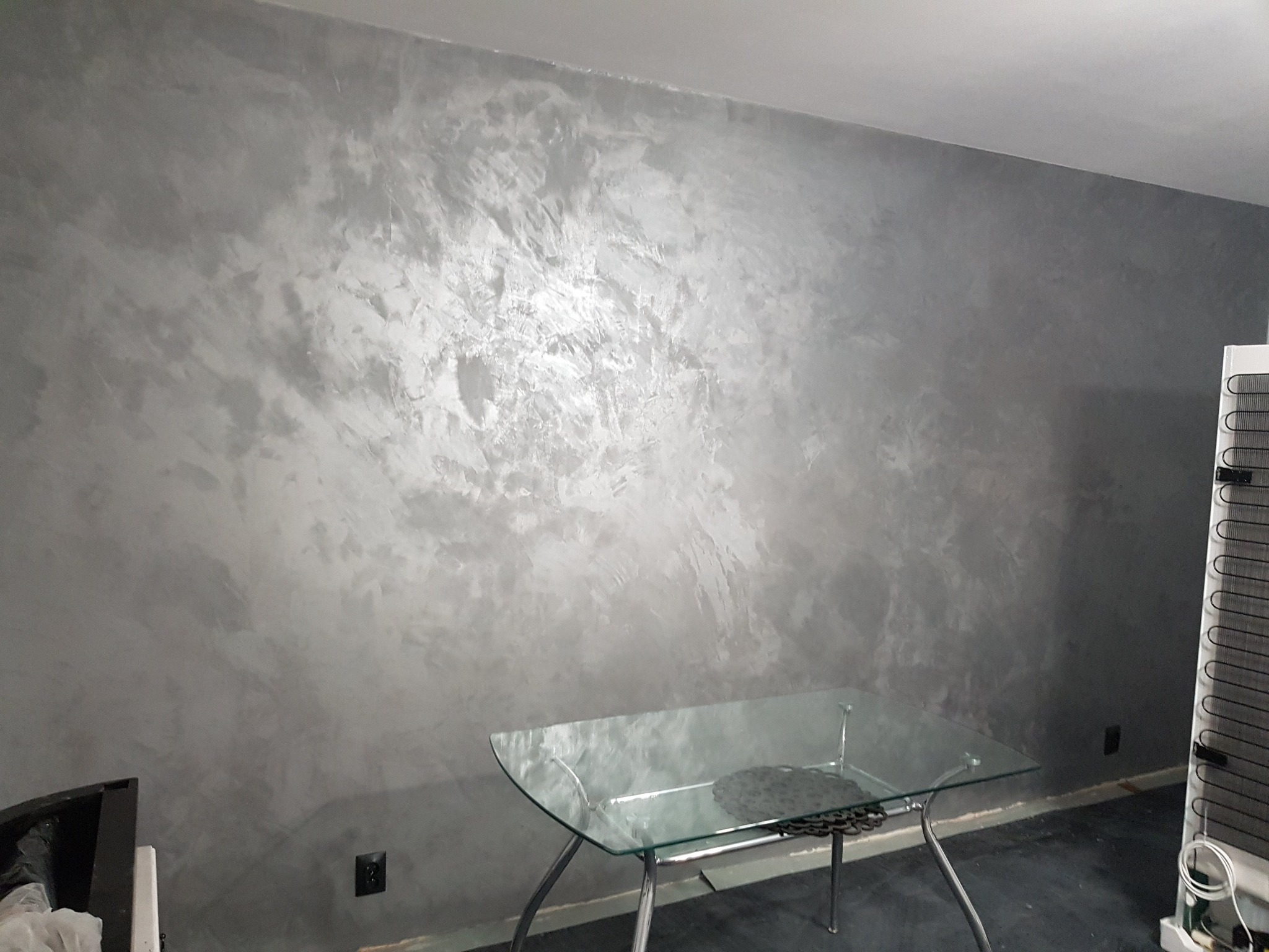 Ściana w salonie wykończona dekoracyjną szarą gładzią w stylu weneckim, z widocznym stołem ze szklanym blatem i fragmentem lodówki.