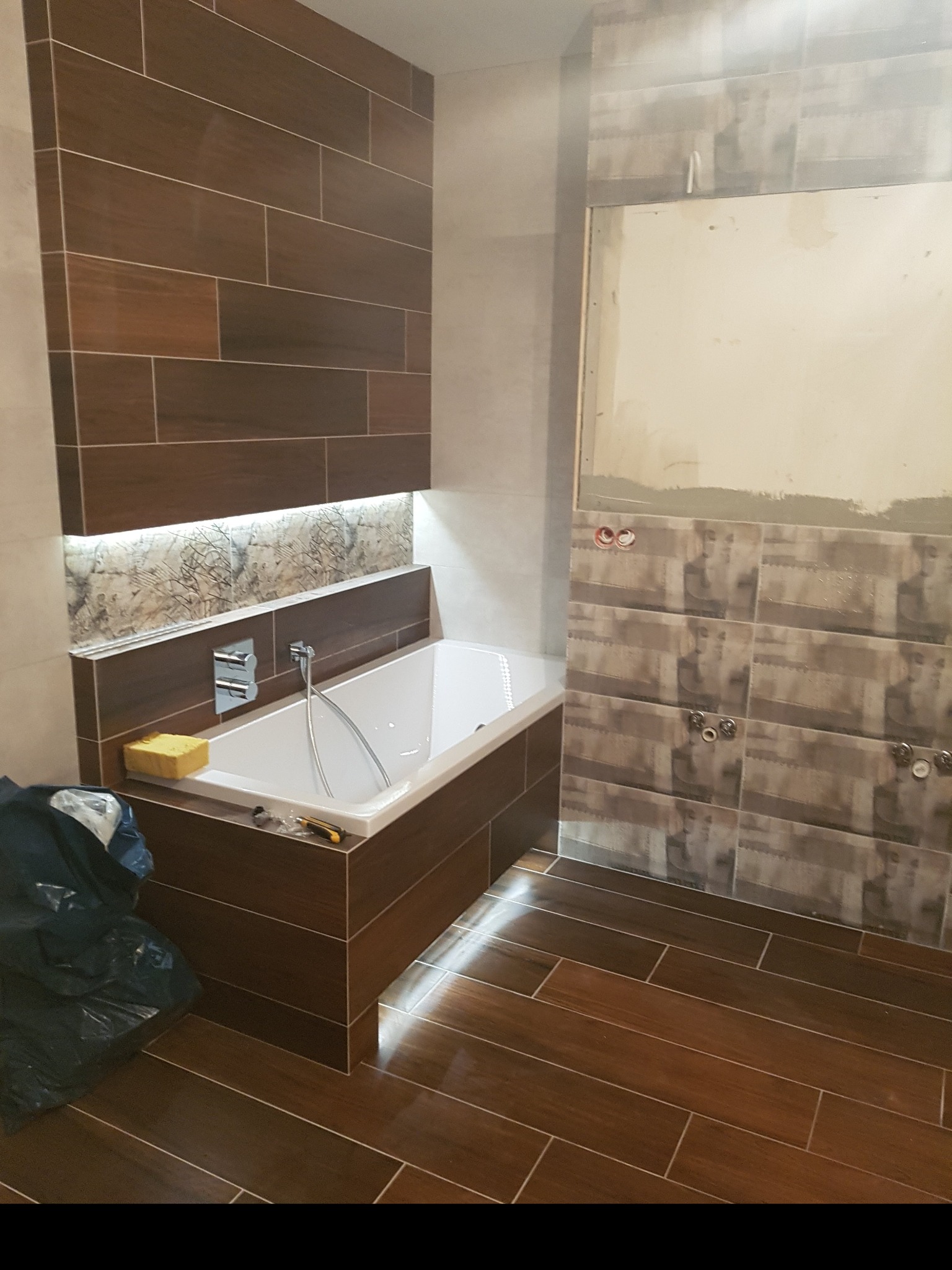 Łazienka w trakcie wykańczania: brązowe płytki imitujące drewno na podłodze i obudowie wanny, częściowo ułożone płytki na ścianie z widocznymi instalacjami, gąbka i narzędzia na wannie, worek...