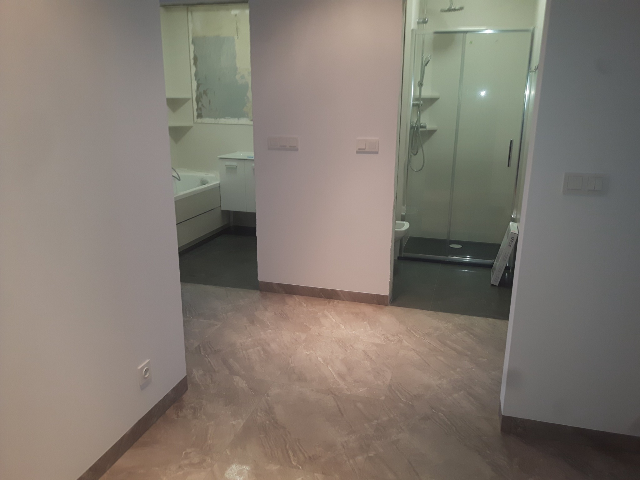 Widok na dwie łazienki z korytarza, widoczna wanna, umywalka, kabina prysznicowa, płytki podłogowe imitujące marmur.