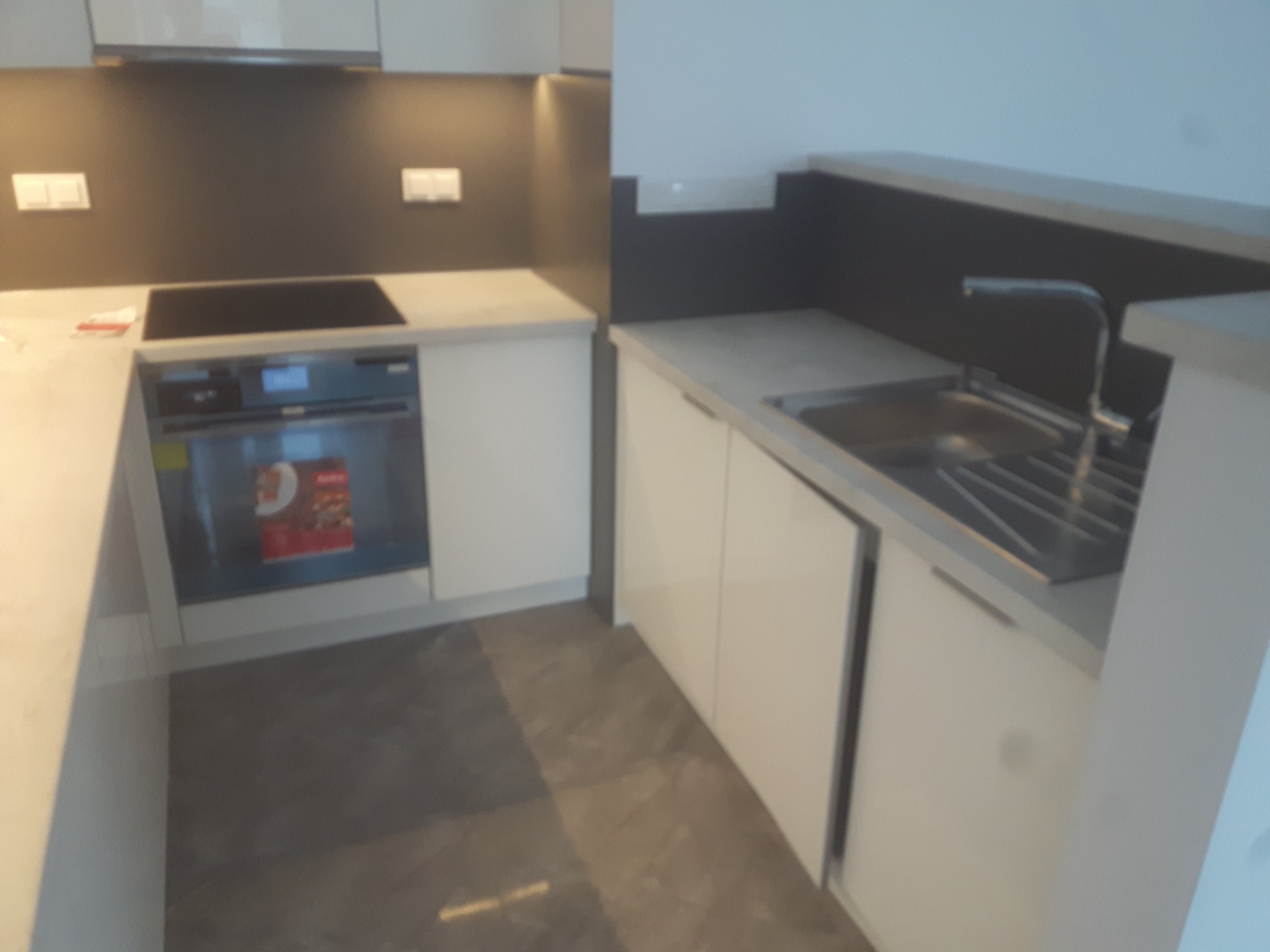 Nowoczesna kuchnia z białymi szafkami, szarą ścianą nad blatem i indukcyjną płytą grzewczą, widoczny zlew z baterią i piekarnik z reklamą produktu.