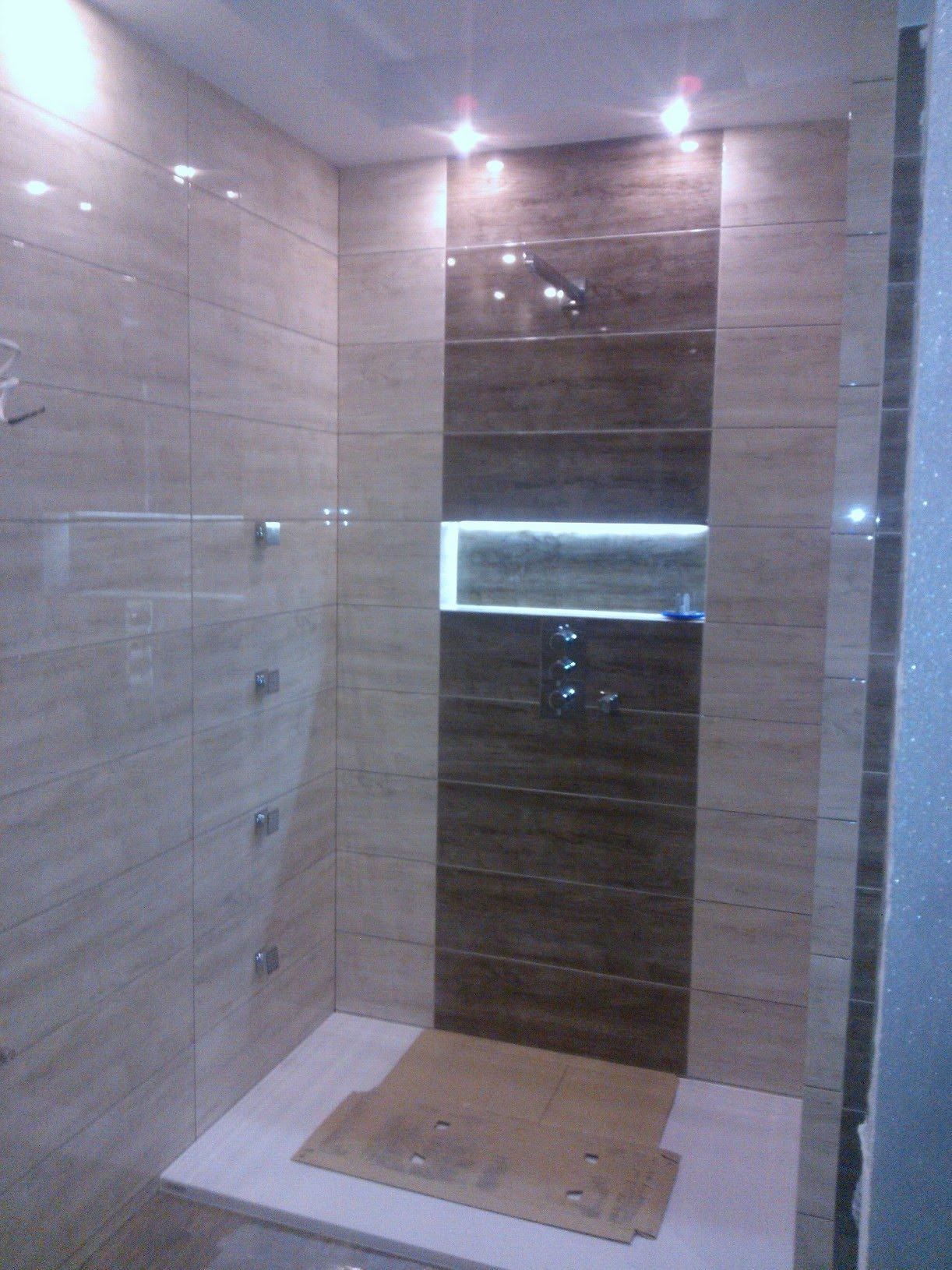 Wykończona kabina prysznicowa z beżowymi i brązowymi płytkami imitującymi drewno, oświetlona wnęka na kosmetyki, chromowana armatura, zabezpieczona podłoga kartonem.