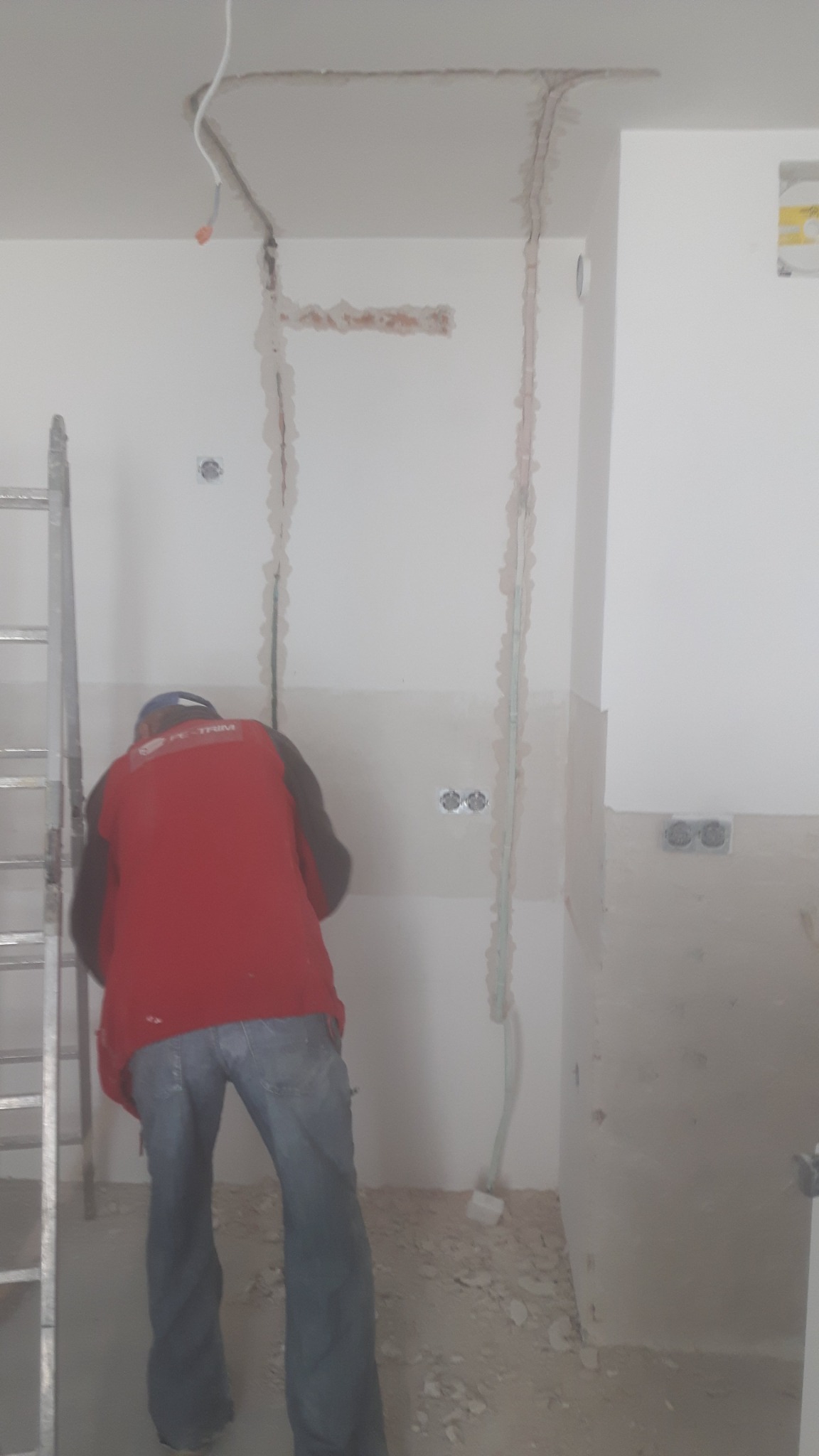 Instalacja elektryczna w bruzdach ściennych podczas prac wykończeniowych, widoczna drabina i fragment sylwetki pracownika w czerwonej koszulce.
