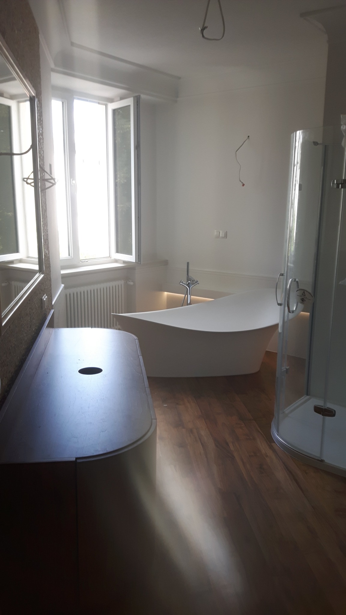 Nowoczesna łazienka z wolnostojącą wanną o asymetrycznym kształcie, drewnianą podłogą i przeszkloną kabiną prysznicową, oświetlona naturalnym światłem z okna.