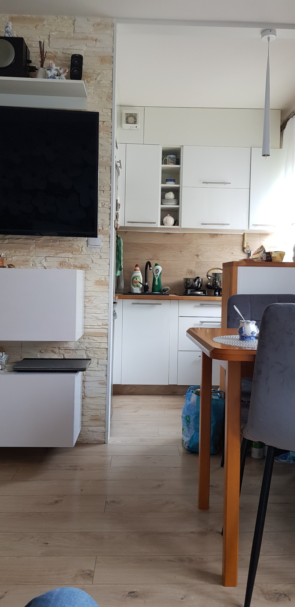 Widok na jasną kuchnię z białymi szafkami i drewnianym blatem, z perspektywy salonu z kamienną ścianą i wiszącym telewizorem.