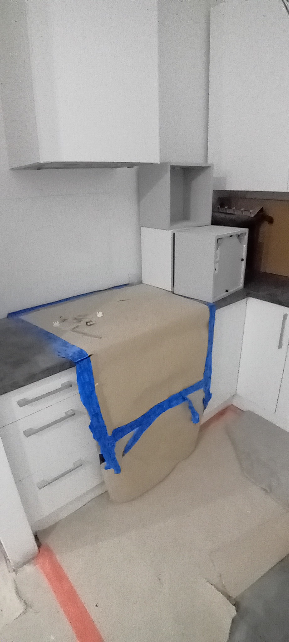 Przygotowanie do prac w kuchni w Płocku: blat zabezpieczony brązowym papierem i niebieską taśmą malarską, widoczne białe szafki kuchenne i fragment podłogi z czerwoną linią.