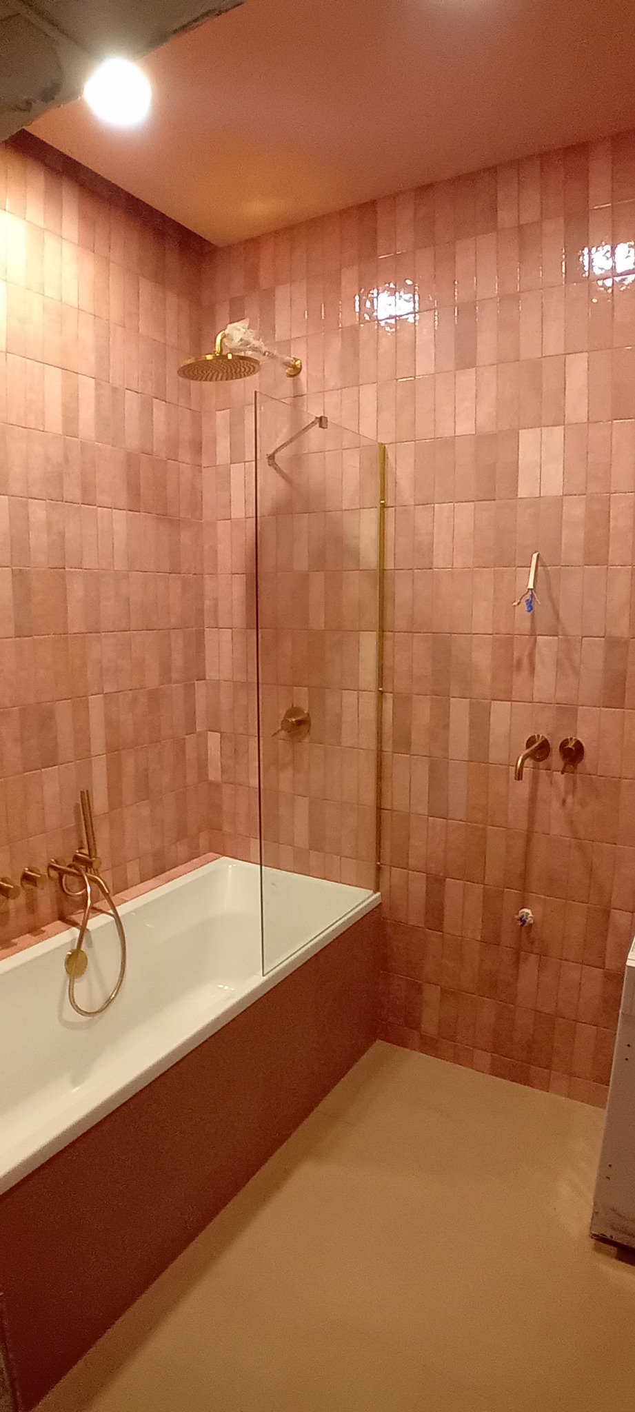 Wnętrze łazienki wykończone w odcieniach różu i złota, z wanną, szklanym parawanem nawannowym i deszczownicą w folii ochronnej.