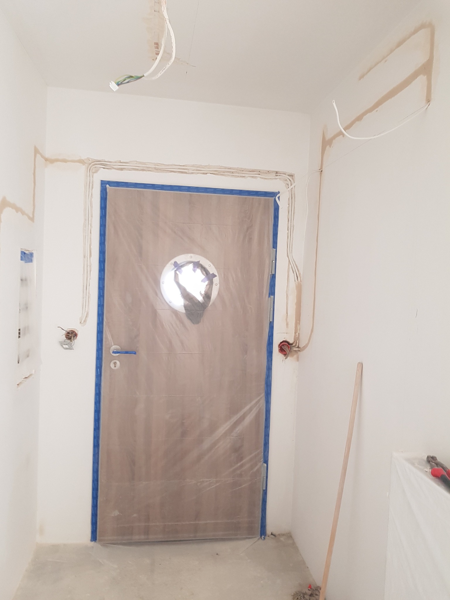 Białe wnętrze w trakcie prac wykończeniowych: zabezpieczone drzwi z okrągłym oknem, widoczne przewody elektryczne i ślady po gładzi na ścianach.