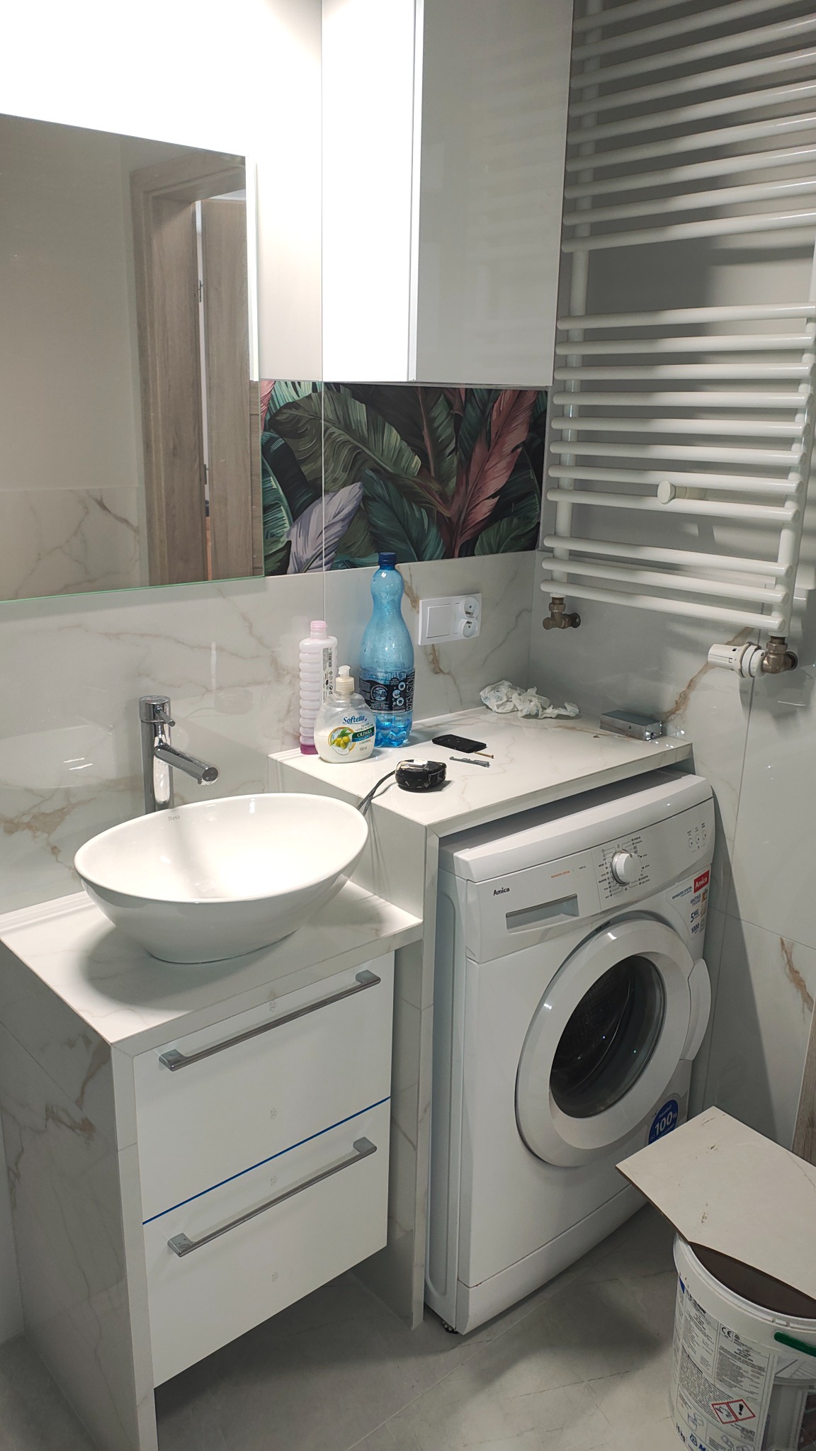 Nowoczesna łazienka z umywalką nablatową, marmurowymi płytkami i motywem tropikalnych liści na ścianie. Biała pralka i grzejnik drabinkowy.