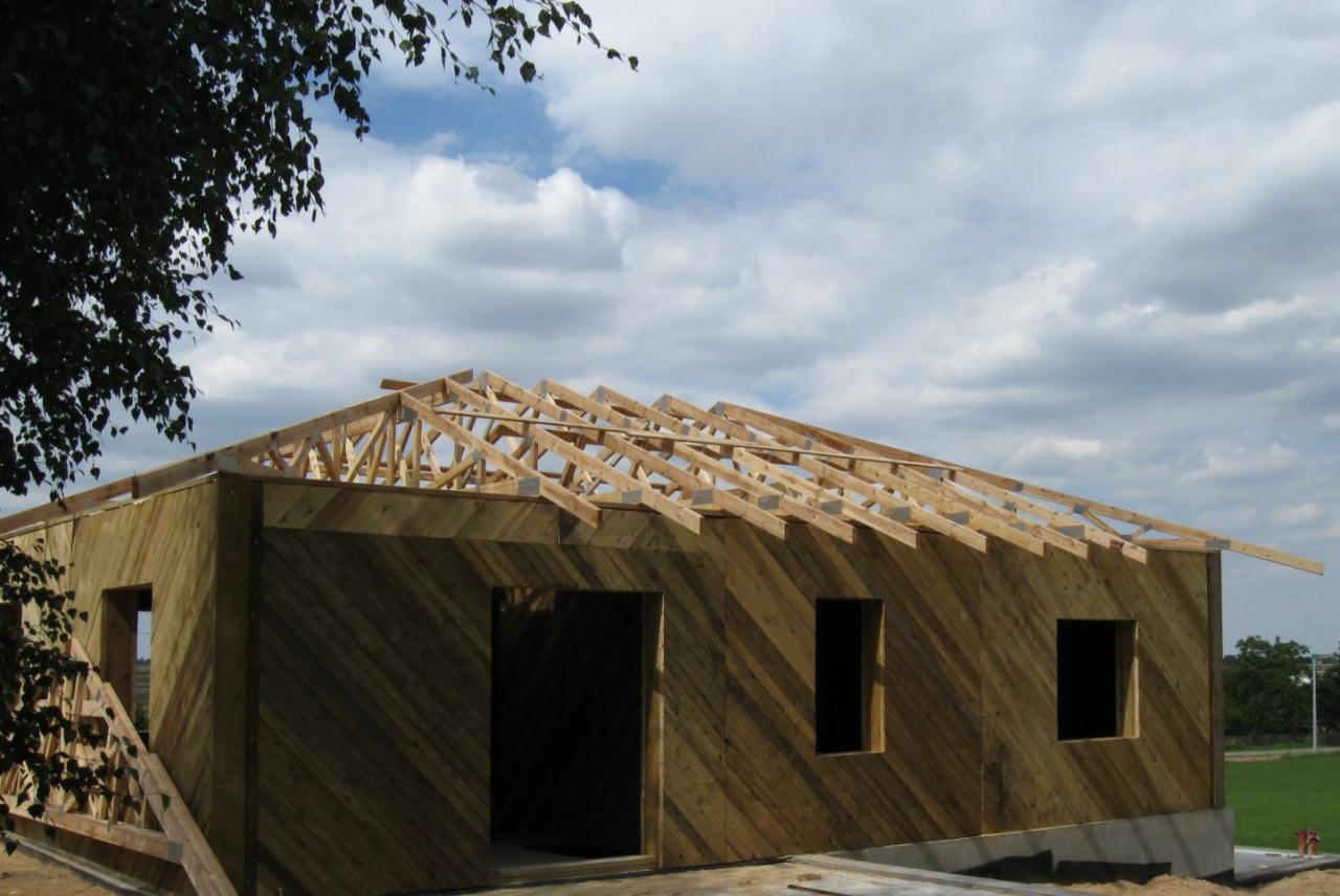 Wznoszenie konstrukcji dachu na budynku z deskami ułożonymi skośnie; widoczne niebo i fragment drzewa.