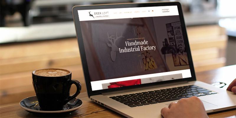 Laptop wyświetla stronę internetową 'Deer Loft Industrial Factory' z kawą latte obok, dłoń użytkownika na touchpadzie.