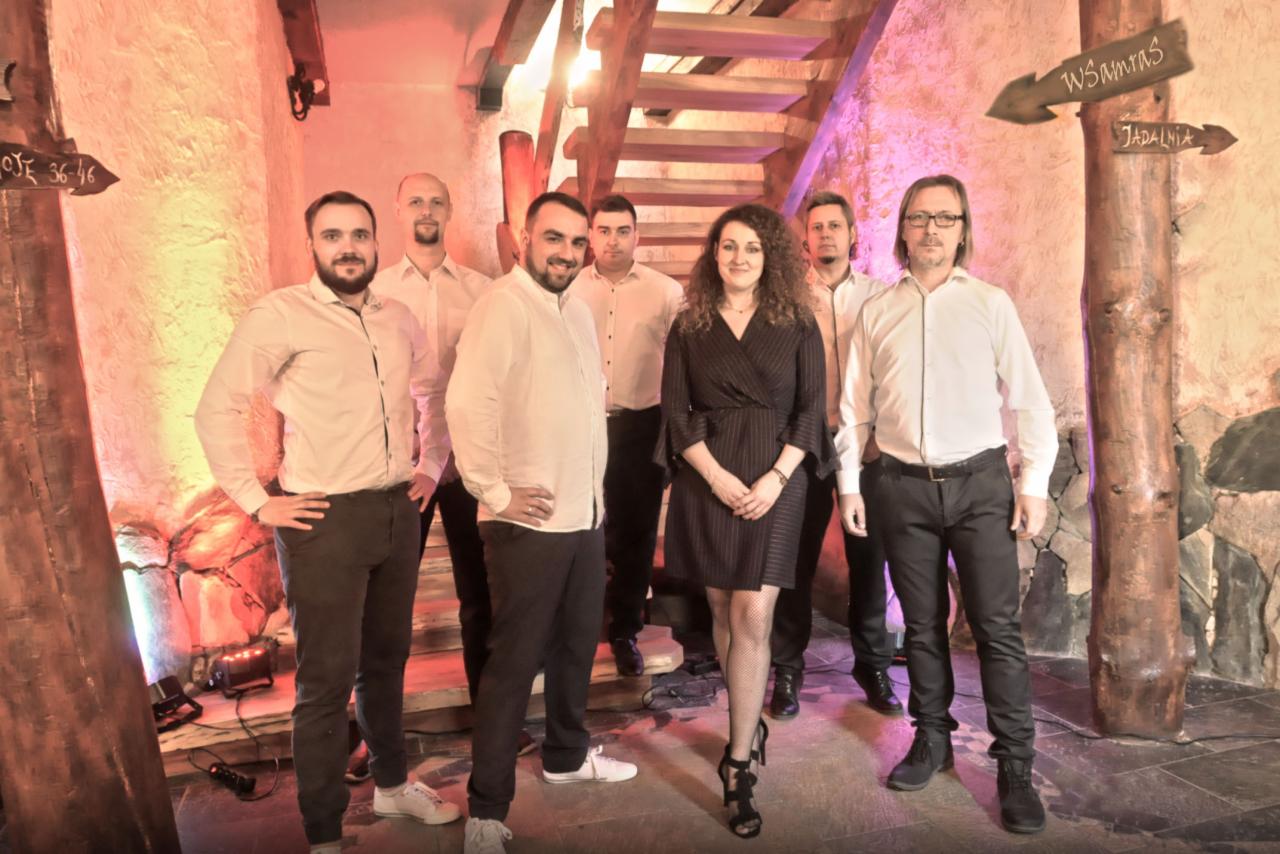 Zespół muzyczny w eleganckich strojach pozuje na tle schodów i dekoracji imitujących drzewo z drogowskazami w rustykalnym wnętrzu sali weselnej.