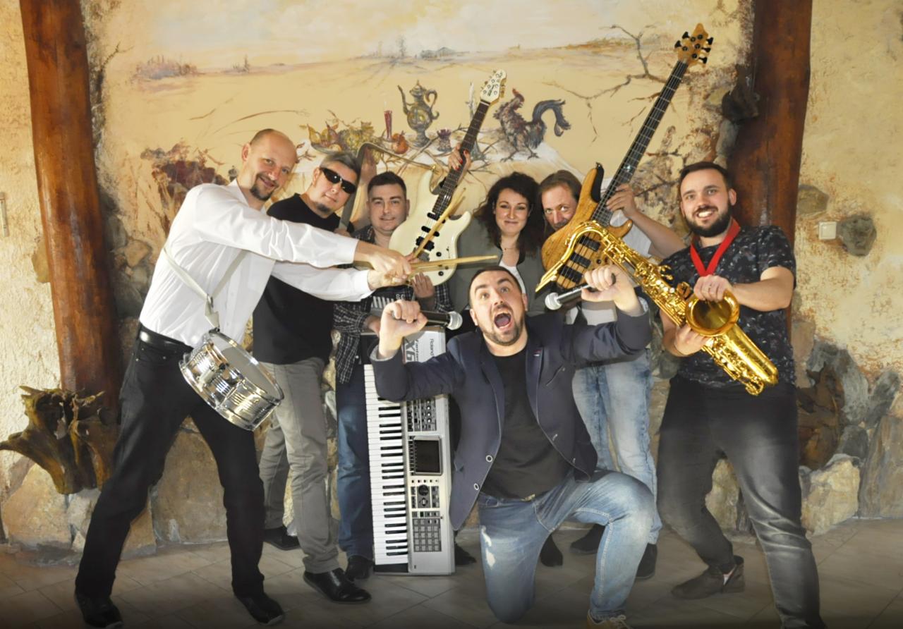 Uśmiechnięta grupa muzyków z instrumentami (saksofon, gitary, keyboard, bębenek) pozuje na tle ściany z malowidłami.