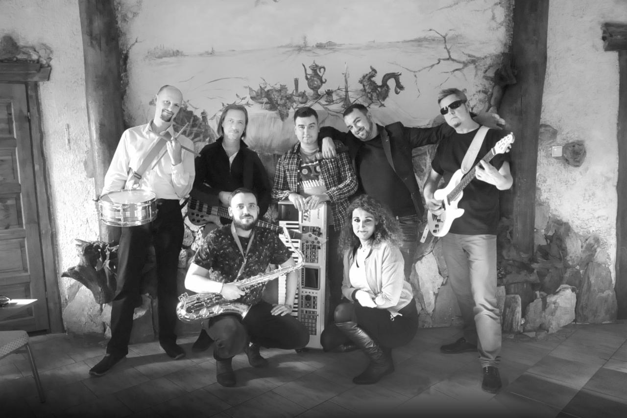 Czarno-białe zdjęcie zespołu muzycznego z saksofonem, perkusją, gitarą i keyboardem, pozującego na tle ściany z malowidłami w rustykalnym stylu.