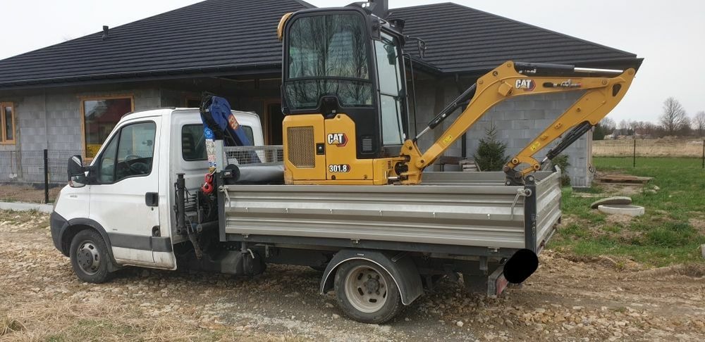 Żółta minikoparka CAT 301.8 załadowana na platformę białego samochodu dostawczego na tle budującego się domu z szarego bloczka.