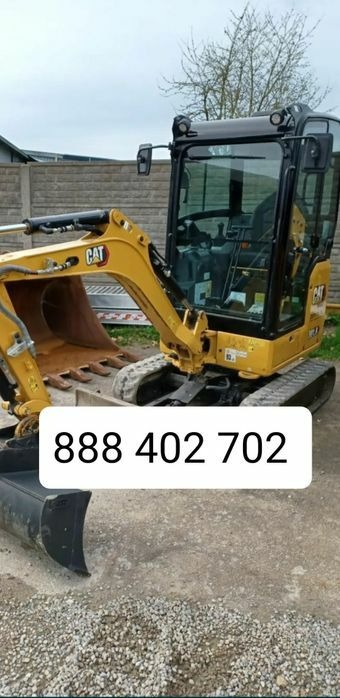 Żółta minikoparka CAT z kabiną, stojąca na utwardzonym podłożu, widoczna łyżka koparki i gąsienice, w tle szary płot.