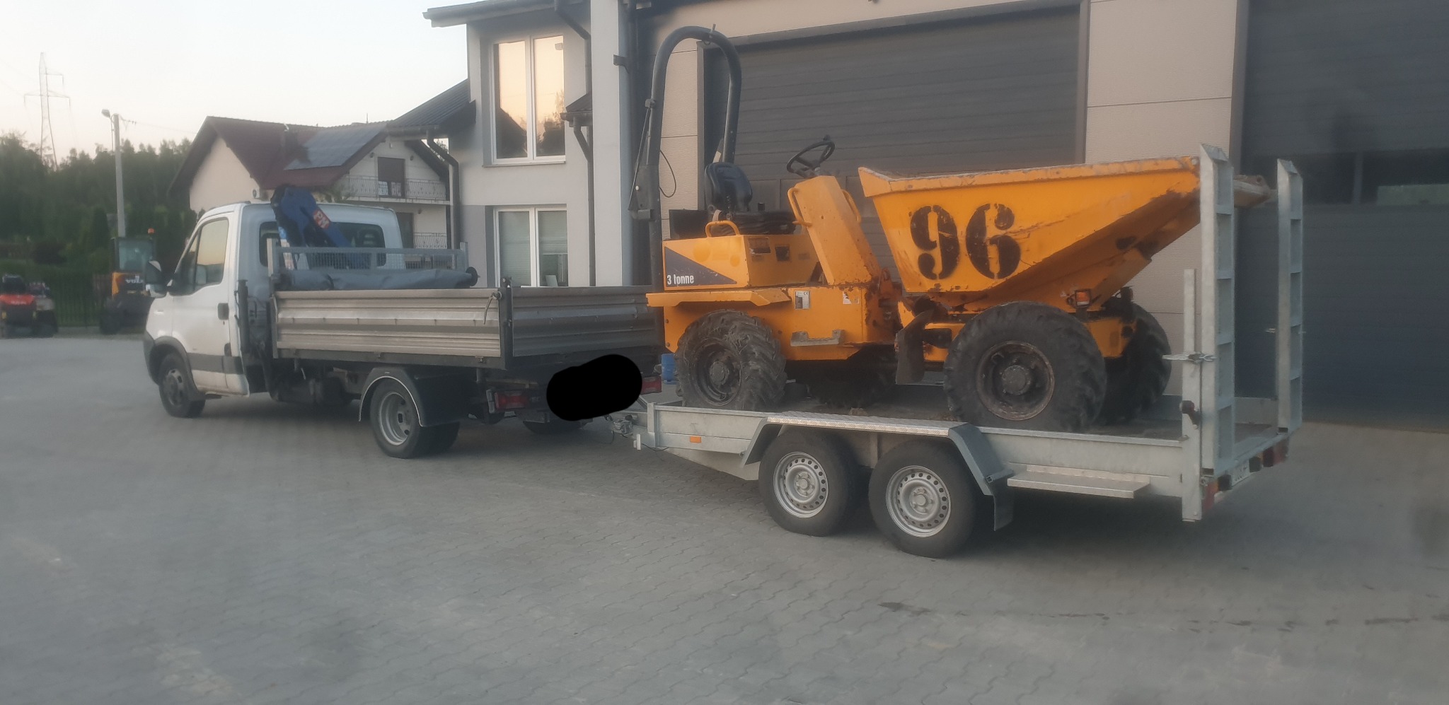 Żółta wywrotka 3-tonowa z numerem 96 na przyczepie, ciągnięta przez biały samochód dostawczy z otwartą skrzynią ładunkową, w tle budynki mieszkalne i garaż.