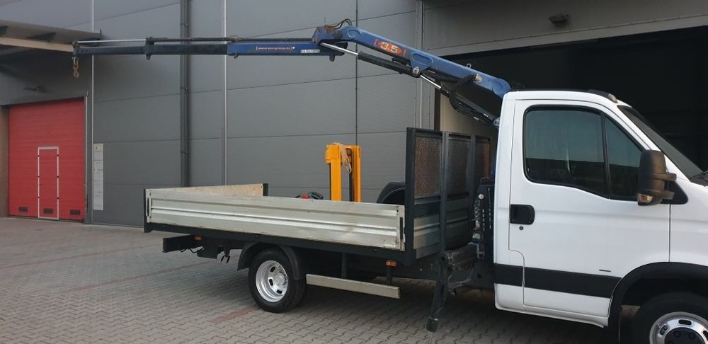 Biały samochód dostawczy z zamontowanym dźwigiem hydraulicznym, zaparkowany na tle szarej hali z czerwoną bramą.