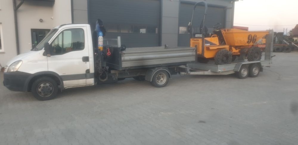 Biały samochód dostawczy z zamontowanym dźwigiem HDS, ciągnący przyczepę z pomarańczową mini wywrotką o numerze 96, zaparkowany na betonowym placu przed budynkiem z szarą bramą.