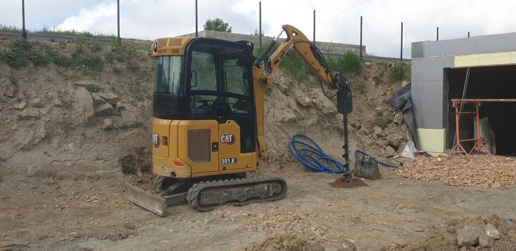 Żółta minikoparka CAT 301.8 z wiertnicą hydrauliczną, niebieskie węże hydrauliczne, na placu budowy z widocznym wykopem i fragmentem budynku w trakcie budowy.