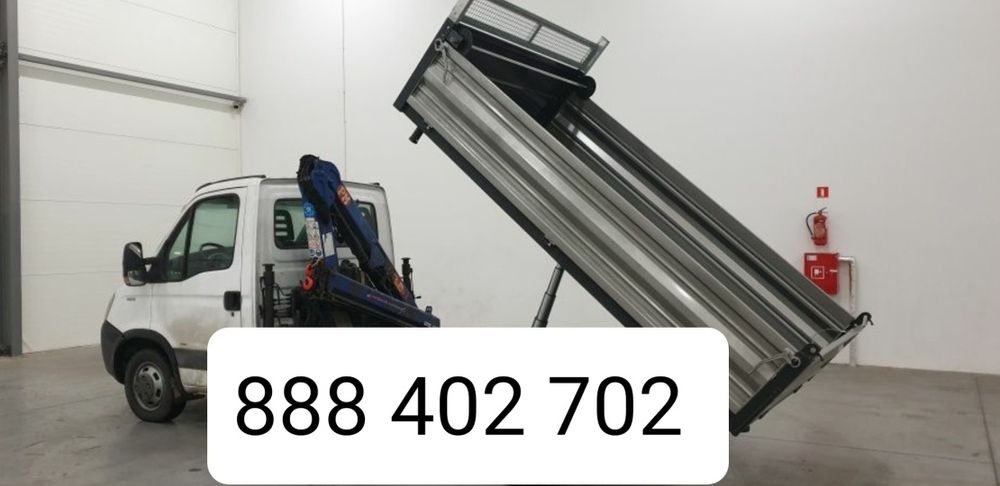 Biały samochód ciężarowy z podniesioną skrzynią i zamontowanym dźwigiem hydraulicznym w jasnym magazynie z białą ścianą i gaśnicą w tle.