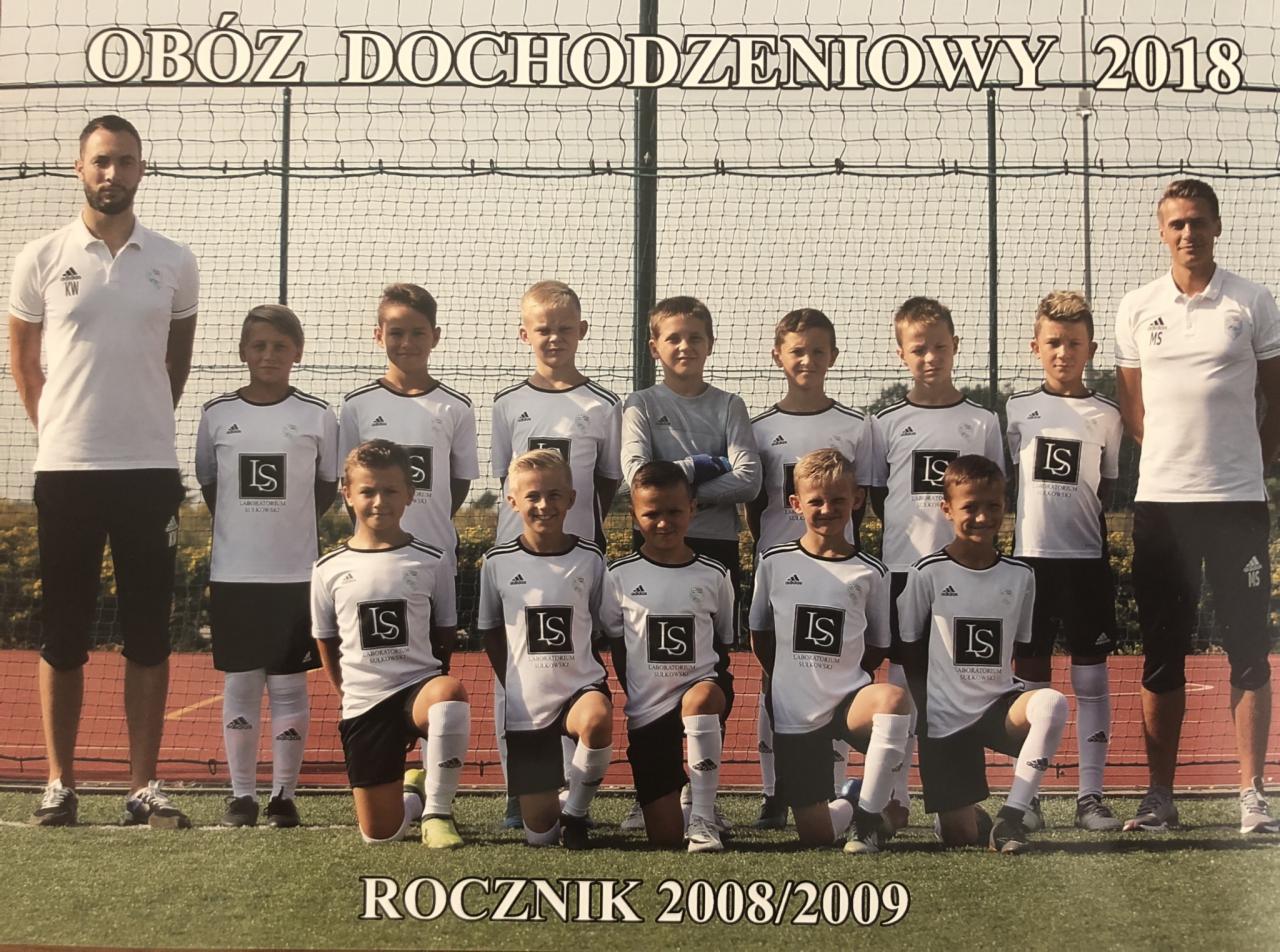 Drużyna piłkarska rocznika 2008/2009 w białych koszulkach z logo sponsora, ustawiona do zdjęcia grupowego na tle siatki i zieleni boiska, z dwoma trenerami po bokach.