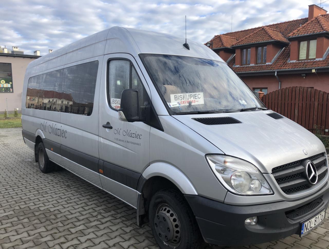 Srebrny bus Mercedes-Benz Sprinter z napisem 'M. Maziec' na boku, zaparkowany na brukowanym placu, z kartką 'Biskupiec Express' za przednią szybą.
