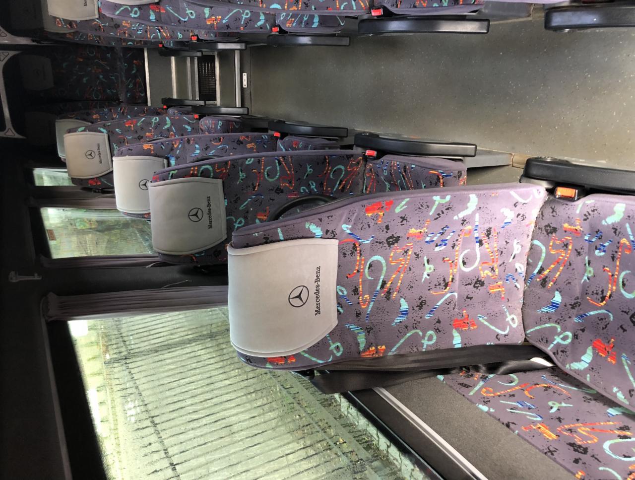 Wnętrze autobusu Mercedes-Benz z rzędem foteli obitych szarą tapicerką w kolorowe wzory, zagłówki z logo producenta.