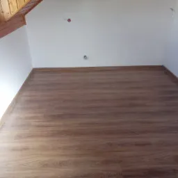 Podłoga z paneli w odcieniach brązu, świeżo ułożona w pokoju na poddaszu ze skośnym sufitem i białymi ścianami. Widoczne gniazdka elektryczne bez osprzętu.