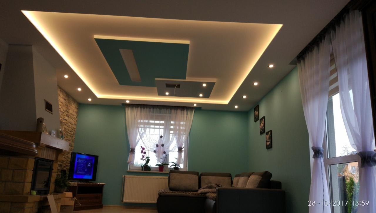 Nowoczesny salon z podwieszanym sufitem z oświetleniem LED, ściany w kolorze turkusowym, kominek z kamienia, okno z białymi firanami.