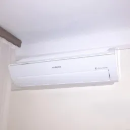 Biała jednostka klimatyzacji Samsung typu split zamontowana pod sufitem w jasnym pomieszczeniu, widoczna z boku, z elementami dekoracyjnymi w tle.