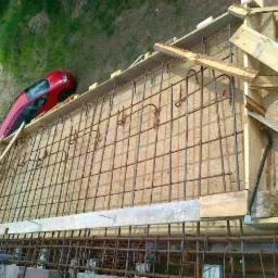 Zbrojenie i szalunek pod wylewkę fundamentu, widoczny fragment muru z cegły i czerwony samochód w tle.