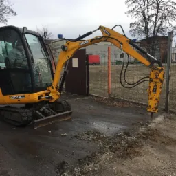 Żółta minikoparka JCB z młotem hydraulicznym krusząca asfalt przy ogrodzeniu z siatki, widoczny operator w kabinie, w tle budynek i zaparkowana ciężarówka.