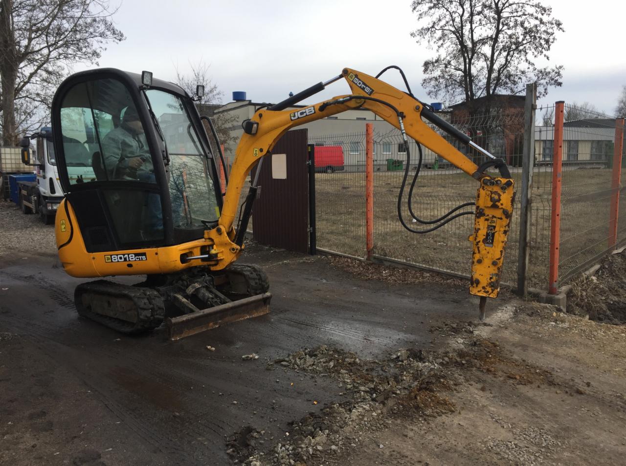 Żółta minikoparka JCB z młotem hydraulicznym krusząca asfalt przy ogrodzeniu z siatki, widoczny operator w kabinie, w tle budynek i zaparkowana ciężarówka.