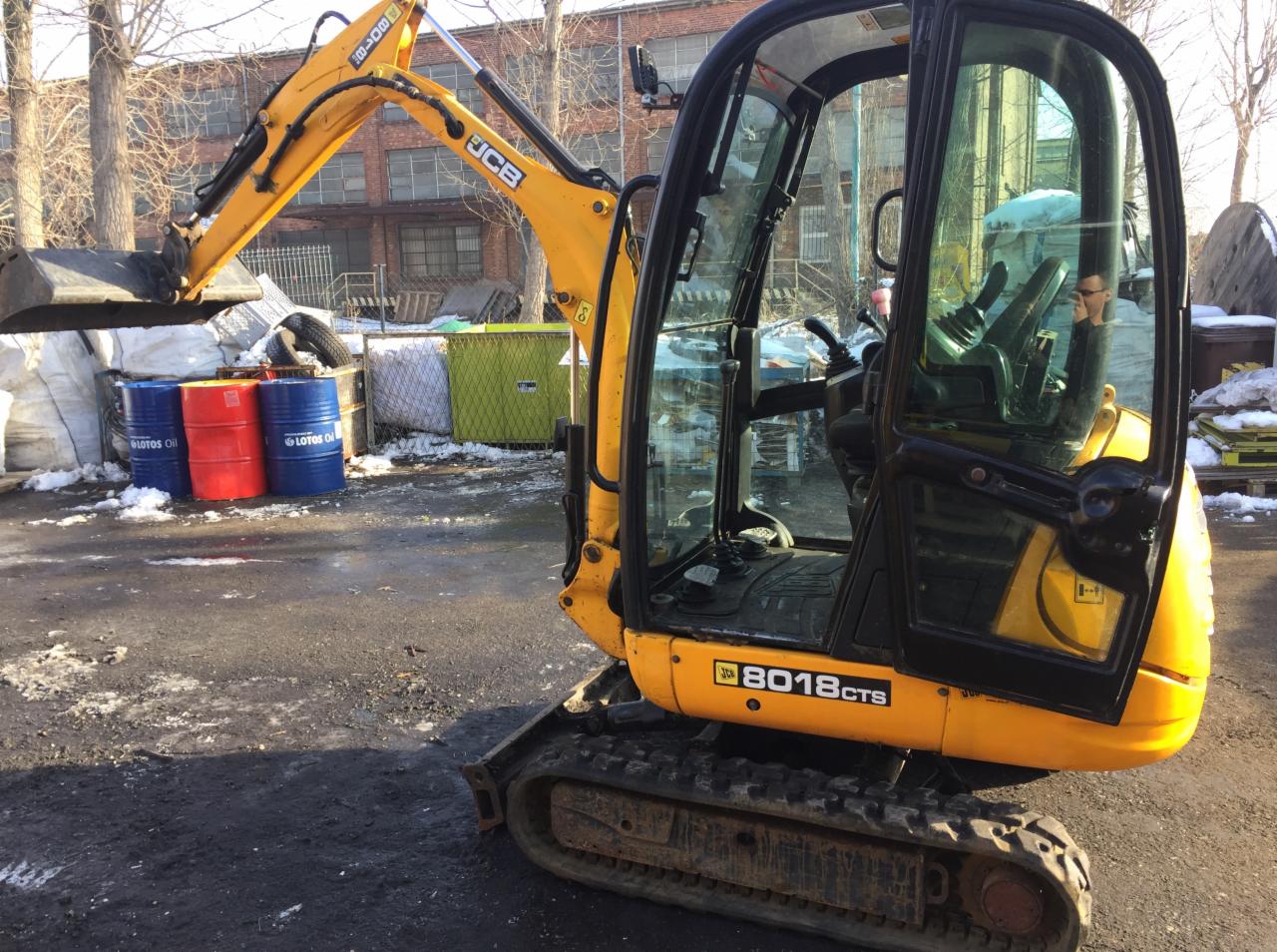Żółta mini koparka JCB 8018 CTS na gąsienicach, z operatorem w kabinie, na placu budowy z beczkami i resztkami śniegu.