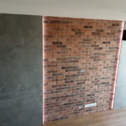 Ściana z cegły klinkierowej z podświetleniem LED, obok szara ściana z płyt imitujących beton, drewniana podłoga, wiszące lampy z czarnymi abażurami.