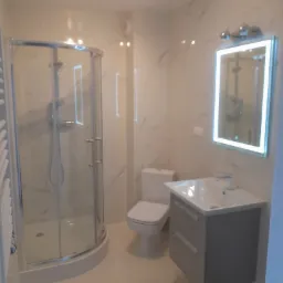 Nowoczesna łazienka z kabiną prysznicową, umywalką z szarą szafką, toaletą i lustrem z oświetleniem LED, wykończona białymi płytkami imitującymi marmur.