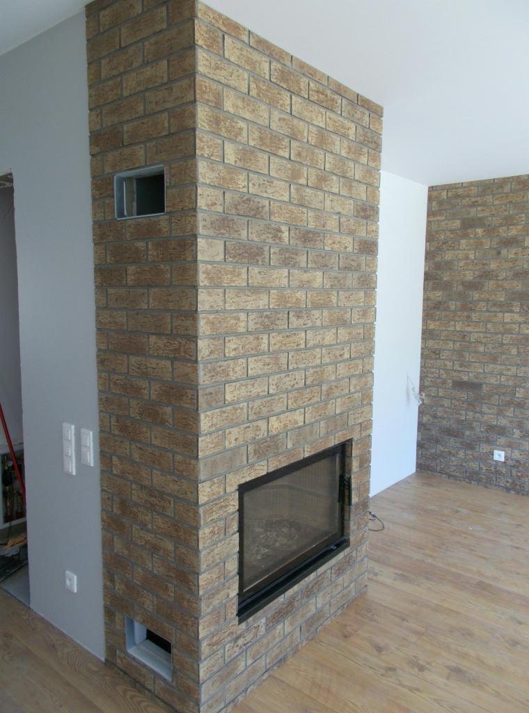 Obudowa kominka z cegły klinkierowej w odcieniach beżu i brązu, z czarnym wkładem kominkowym i kratkami wentylacyjnymi, na tle drewnianej podłogi i fragmentu ściany z podobną okładziną.