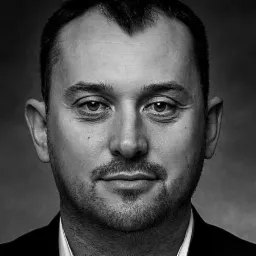 Portret czarno-biały mężczyzny z krótką brodą, lekko uśmiechniętego, w ciemnym garniturze, na rozmytym tle.