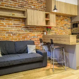 Salon z aneksem kuchennym w stylu loftowym: ceglana ściana, szara sofa, drewniane meble kuchenne i podłoga, nowoczesne oświetlenie.